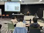A presenter at a lectern explaining a slide on 4D LiDAR for robot dogs, with a robot dog lying on the floor near the audience.
