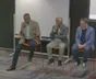 Three panellists seated on tall stools, one speaking into a microphone.