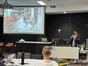 A speaker with a microphone beside a projected photo of a four-legged robot walking through a construction site.